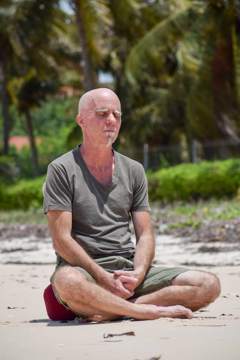 Mindfulness meditation teacher Gerry Rickard sits calmly on a quiet beach in Mozambique during a meditation session, surrounded by soft ocean waves, pale sand, and warm coastal light