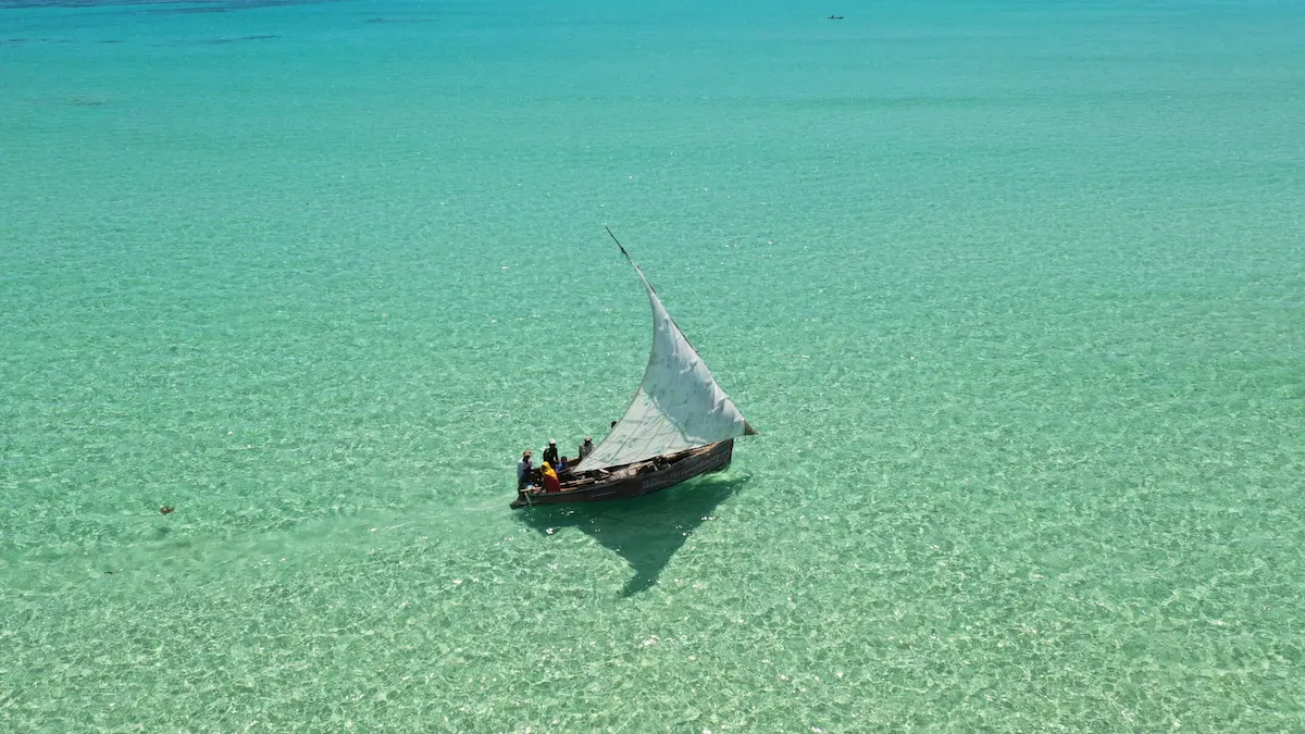 A traditional dhow boat sails across the crystal-clear turquoise waters off the coast of Mozambique under bright blue skies, capturing the peaceful beauty and coastal spirit for a retreat in Mozambique